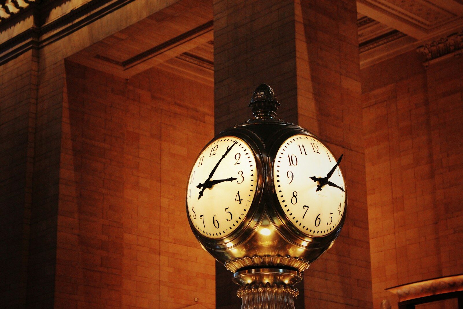 A four-faced brass station clock in a grand hall, two faces showing their own times