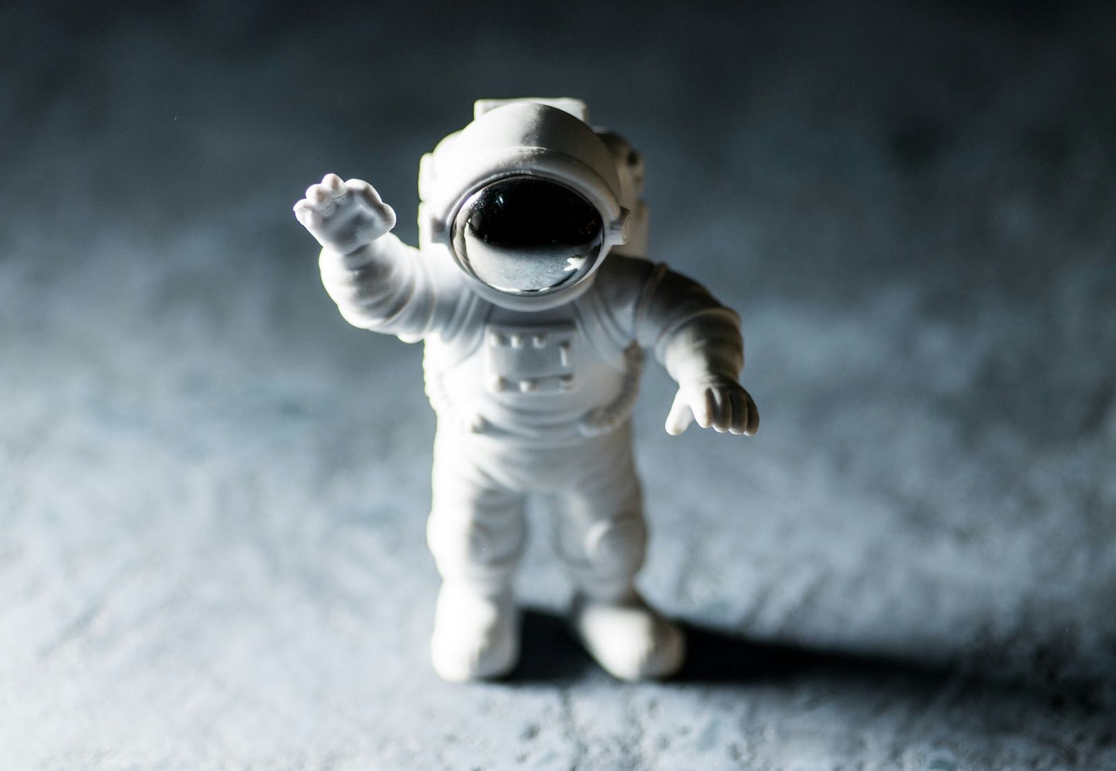 A small white toy astronaut standing on grey moon dust, one arm raised in a wave, casting a long shadow