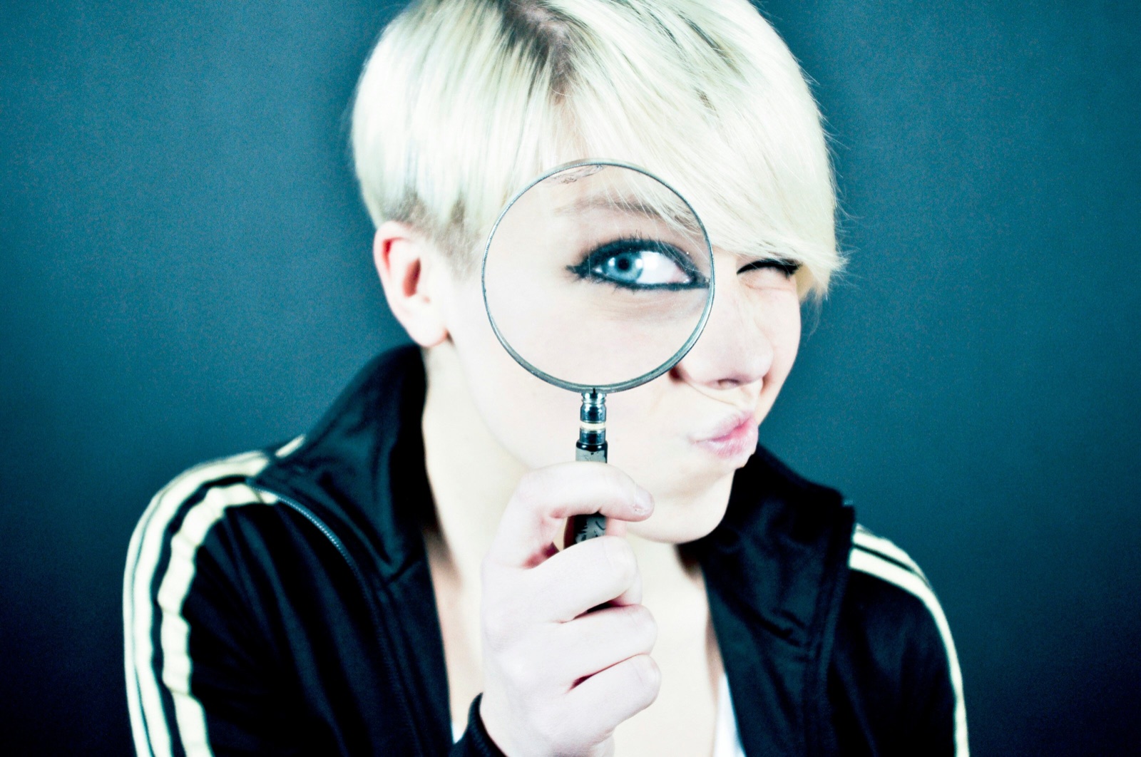 A woman with short platinum-blonde hair and a black track jacket holds a magnifying glass to her face against a dark blue-grey background. One eye looks huge through the lens, and her other eye is closed.